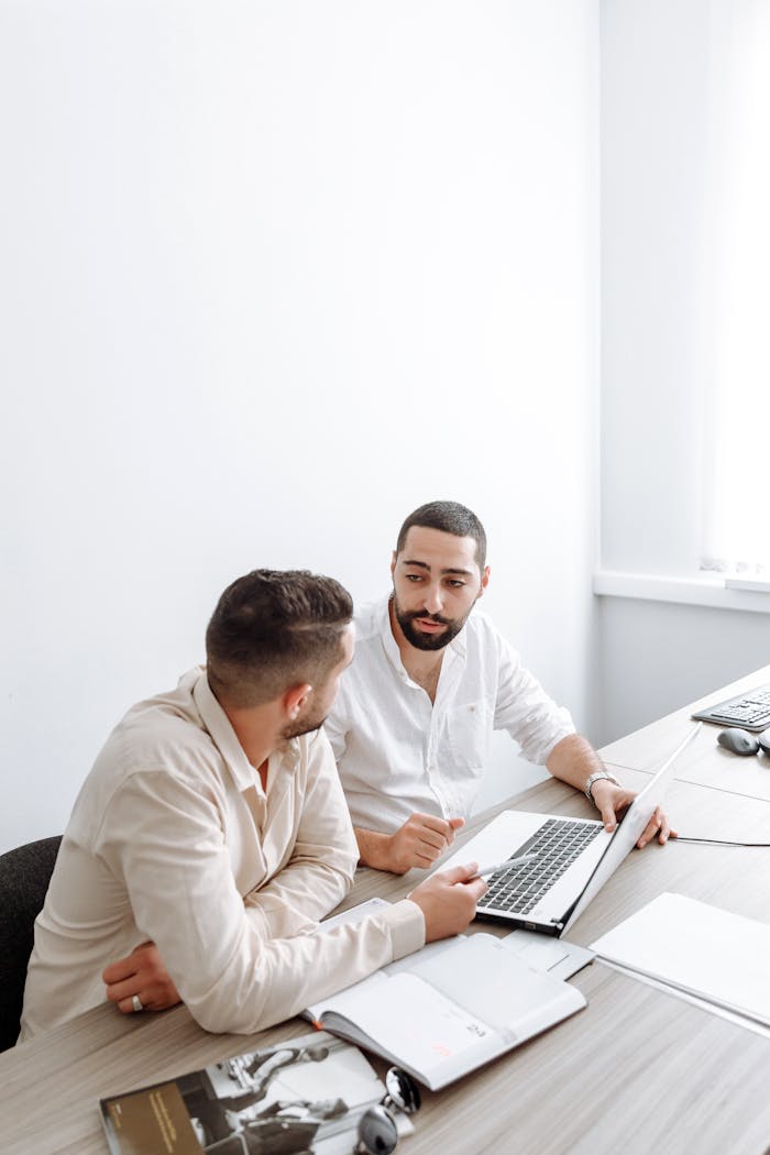 Two businessmen in white shirts engaged in a discussion over a laptop in a bright office setting.